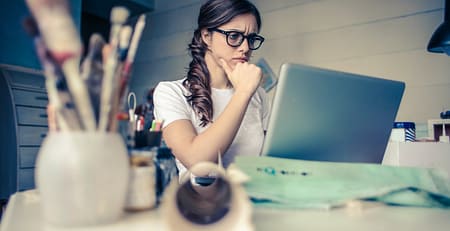 A woman in glasses is sitting at a desk with a laptop, seeking academic assistance.