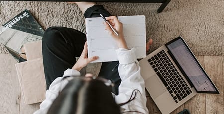A woman at a desk, writing in a notebook and using a laptop is looking for strategies to start and finish her essay on a perfect note.